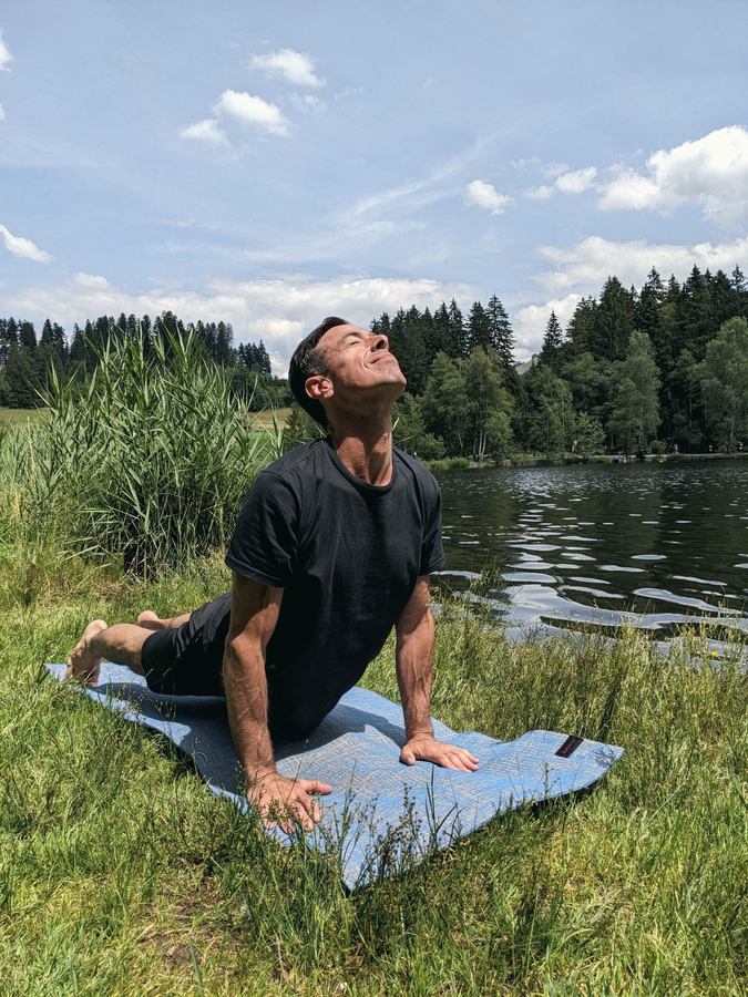 Gustavo in a gentle backbend on a mat by the lake, face turned to the sun