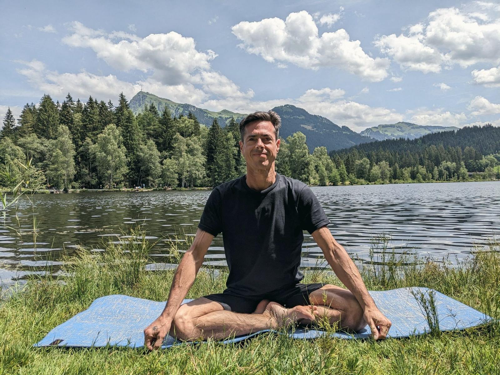 Gustavo sitting cross-legged on a yoga mat by a mountain lake, smiling calmly